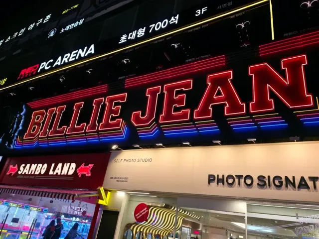 A giant neon sign on a building front at night, with lit shops below it on the street
