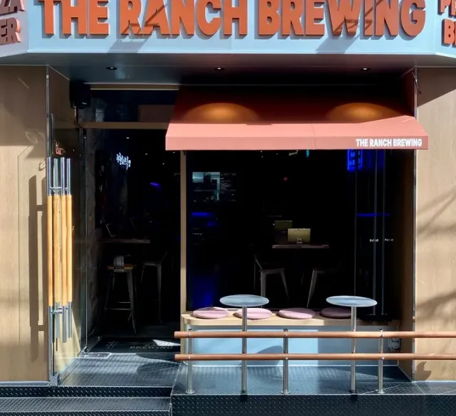 A front with large orange letters, a pink awning and round tables at the open entrance
