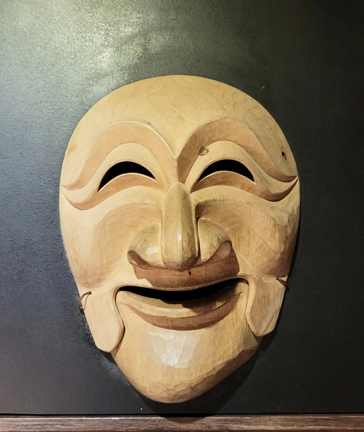 A smiling wooden mask with half closed eyes, lit against a dark background