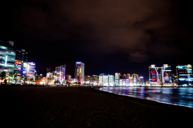A dark strip of beach facing a row of neon lit seafront buildings reflected in the water at night