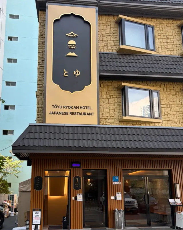 A building front with a large vertical sign, a dark tiled roof and a ground floor clad in wooden boards